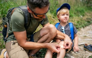 Mature father with small son hiking outdoors in summer nature, putting plaster on knee.