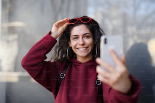 Un ritratto di giovane donna con smartphone in piedi all'aperto sulla strada in città, scattando selfie.