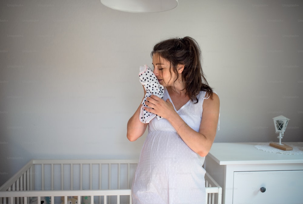 Front view portrait of pregnant woman indoors at home, holding girls bodysuit.