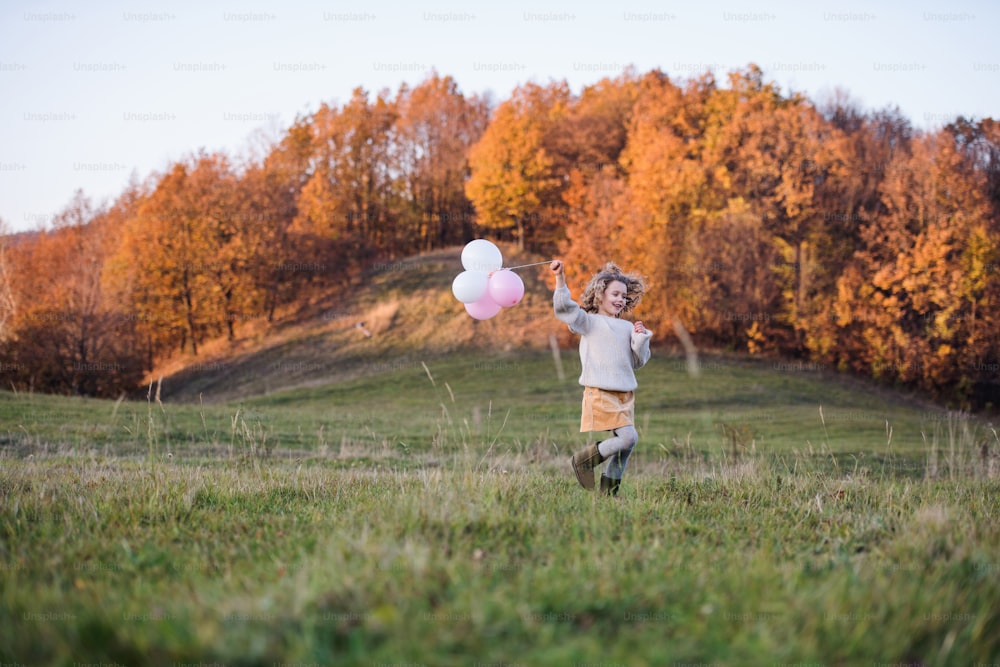 Cheerful small girl with balloons running on meadow in autumn nature.