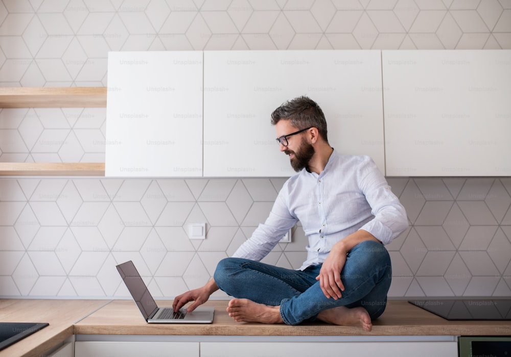 Un homme mûr avec un ordinateur portable assis dans la cuisine dans une nouvelle maison non meublée. Un nouveau concept de maison.