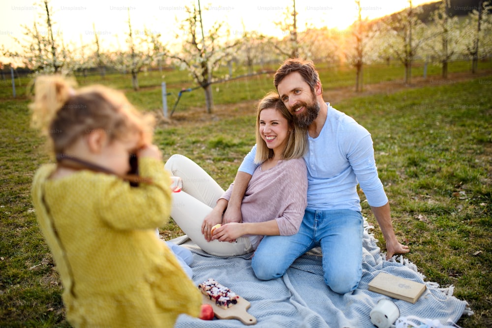 Small girl with camera taking photograph of family on picnic outdoors in spring nature at sunset.