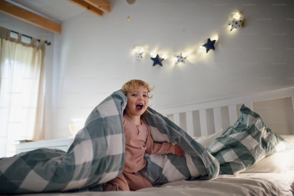 Small toddler girl playing and hiding on bed indoors at home, having fun.