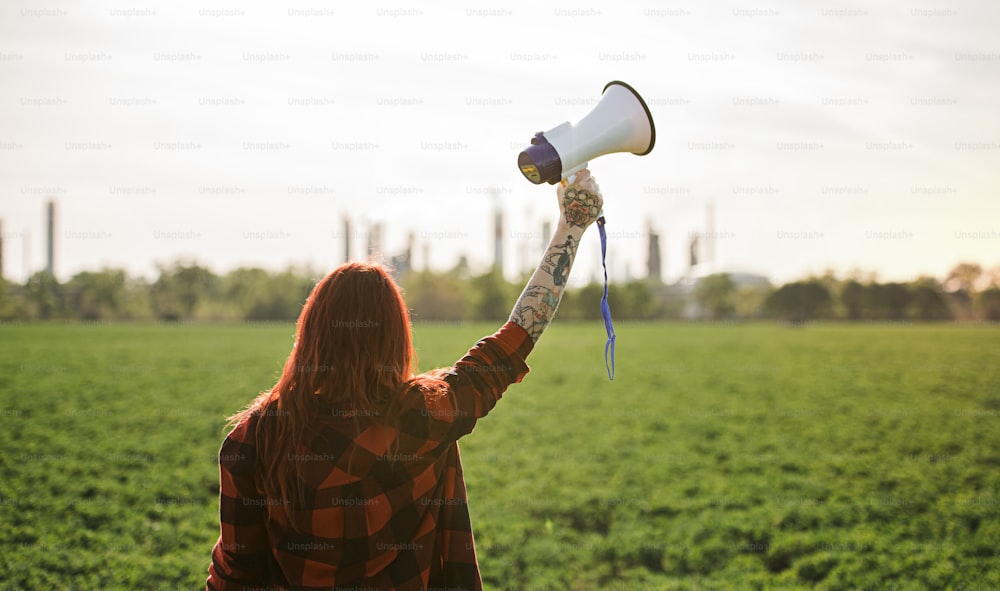 Vista trasera de una joven activista con megáfono de pie al aire libre junto a una refinería de petróleo, protestando.
