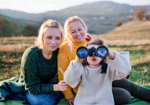 쌍안경을 사용하여 가을 자연 속에서 산책하는 어머니와 할머니와 함께 있는 작은 소녀의 초상화.