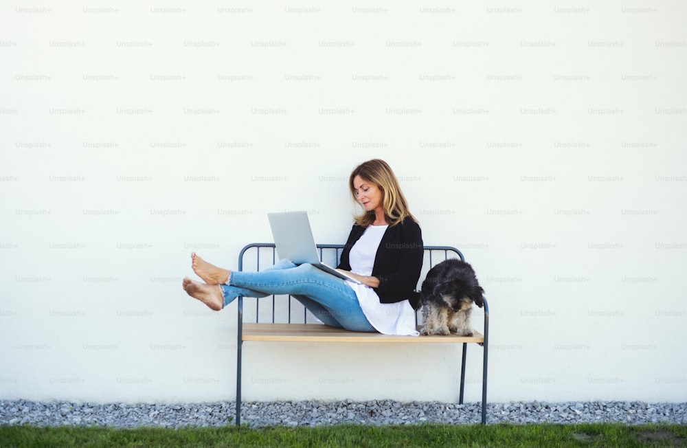 Glückliche reife Frau mit Hund, die im Home Office draußen auf der Bank arbeitet und Laptop benutzt.