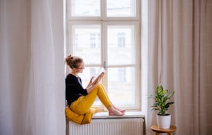 Eine junge, glückliche Studentin mit einem Buch, das zu Hause auf der Fensterbank sitzt und studiert.
