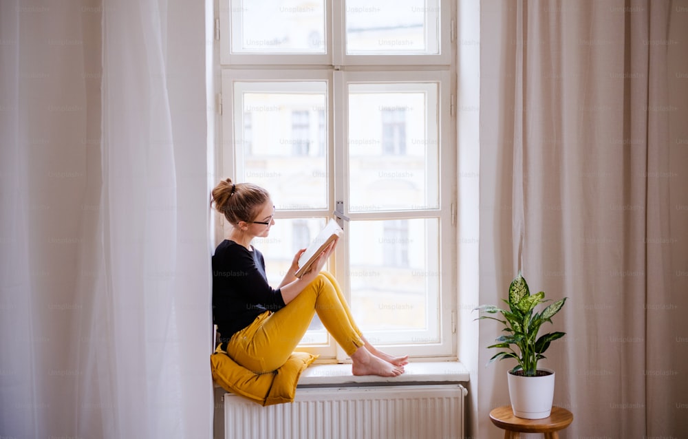 Una joven estudiante universitaria feliz con un libro sentado en el alféizar de la ventana de su casa, estudiando.