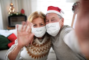 Front view of happy senior couple with face masks indoors at home at Christmas, taking selfie.