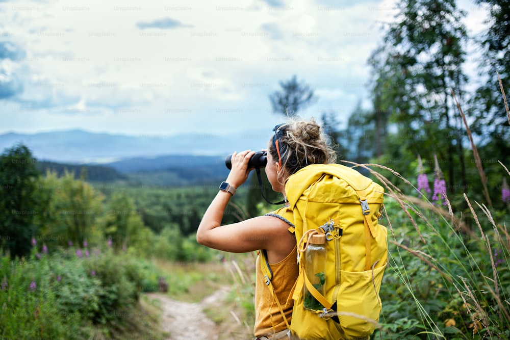 Vista trasera de una joven excursionista con mochila en un viaje de senderismo en la naturaleza, usando binoculares.