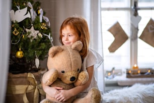 Portrait of small girl standing indoors at home at Christmas, hugging teddy bear.