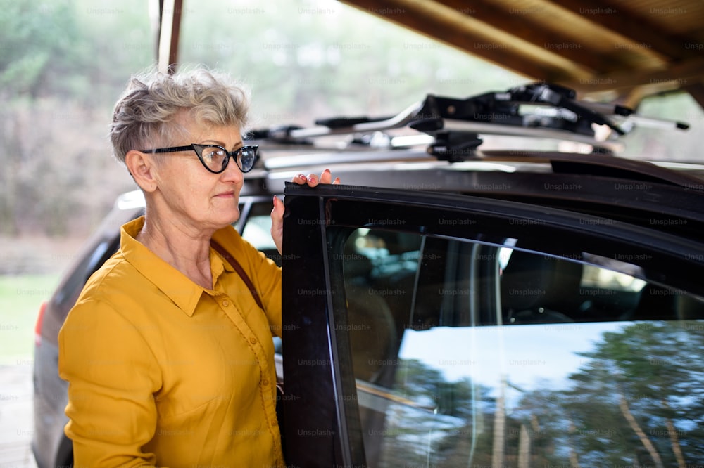 Senior woman with glasses standing outdoors by car by house. Copy space.