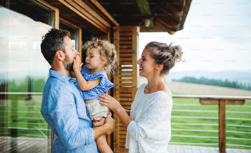 Family with small daughter standing on patio of wooden cabin, holiday in nature concept.