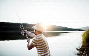 夕暮れ時に湖畔で釣りをする帽子をかぶった成熟した男性が竿を持って釣りをしている。スペースをコピーします。