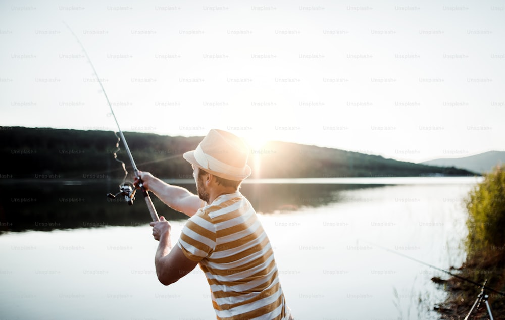 A mature man with a hat fishing by a lake at sunset, holding a rod. Copy space.