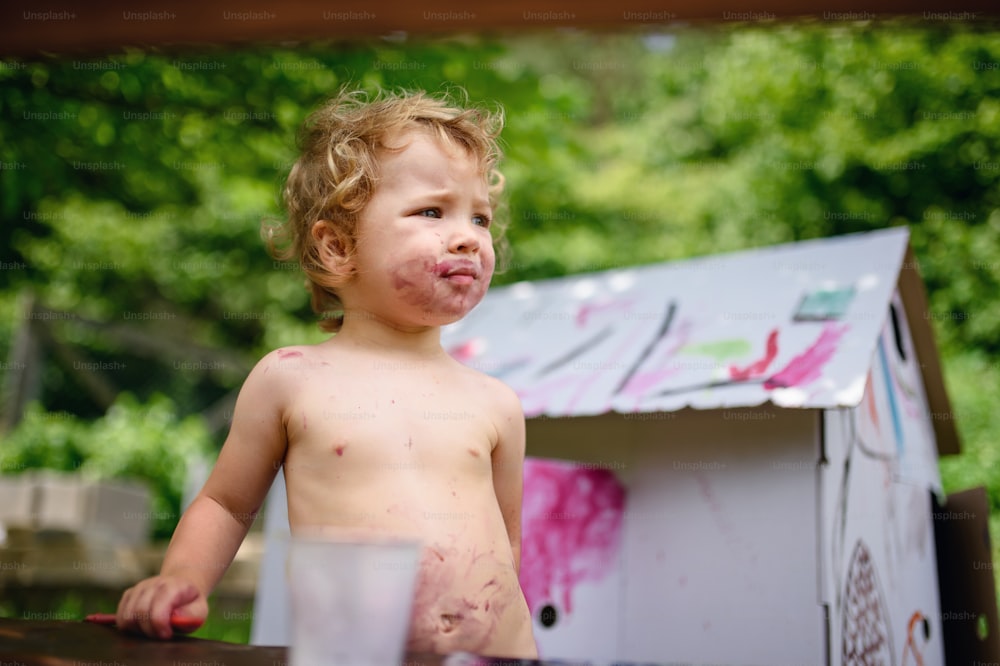 Happy topless small blond girl painting paper house outdoors in summer.