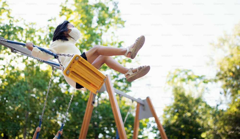 Petite fille avec un masque facial sur une balançoire sur une aire de jeux à l’extérieur en ville, concept de coronavirus.