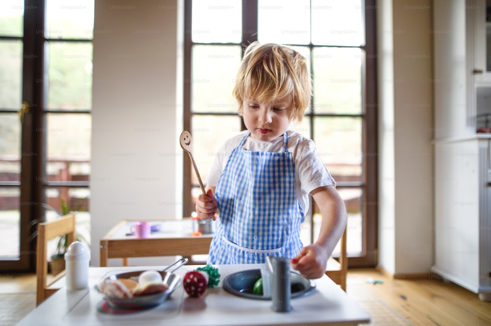 Menino pequeno feliz com avental brincando dentro de casa com cozinha de brinquedo em casa.