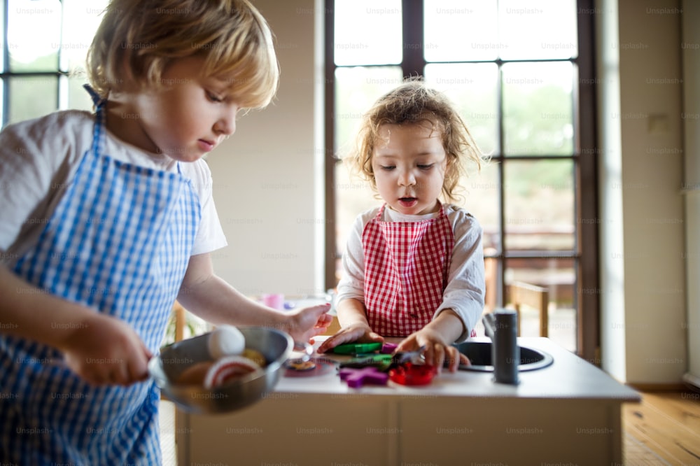 Menino pequeno feliz e menina com avental jogando dentro de casa com cozinha de brinquedo em casa.