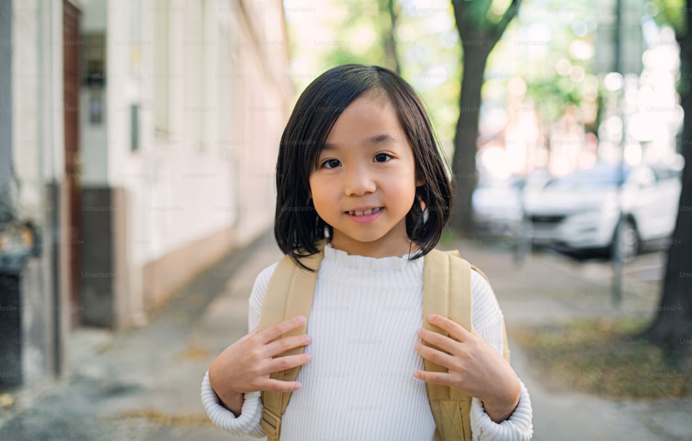 Portrait d’une petite fille japonaise avec un sac à dos marchant à l’extérieur en ville, regardant la caméra.