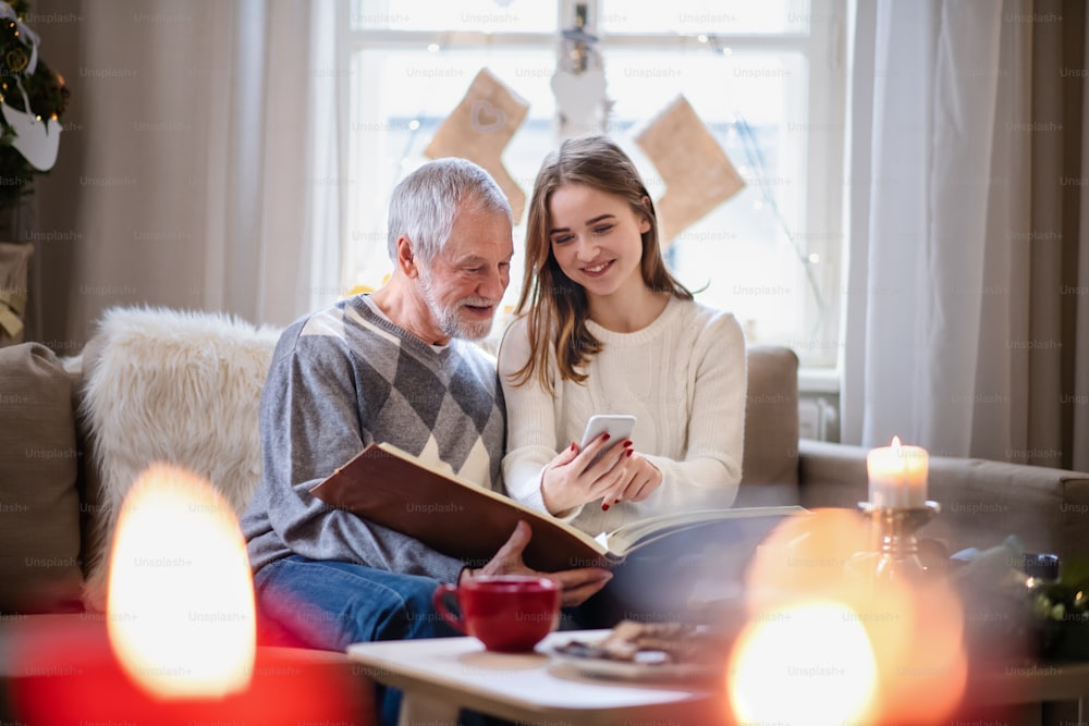 Portrait de jeune femme avec grand-père à l’intérieur à la maison à Noël, à l’aide d’un smartphone.