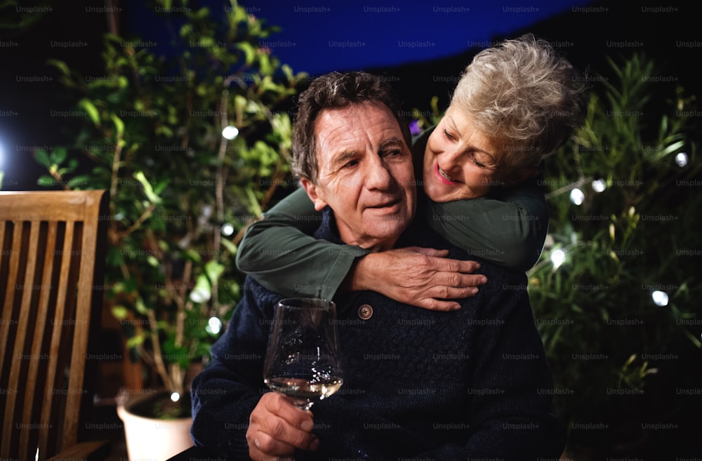 Portrait of senior couple with wine in the evening on terrace, hugging.