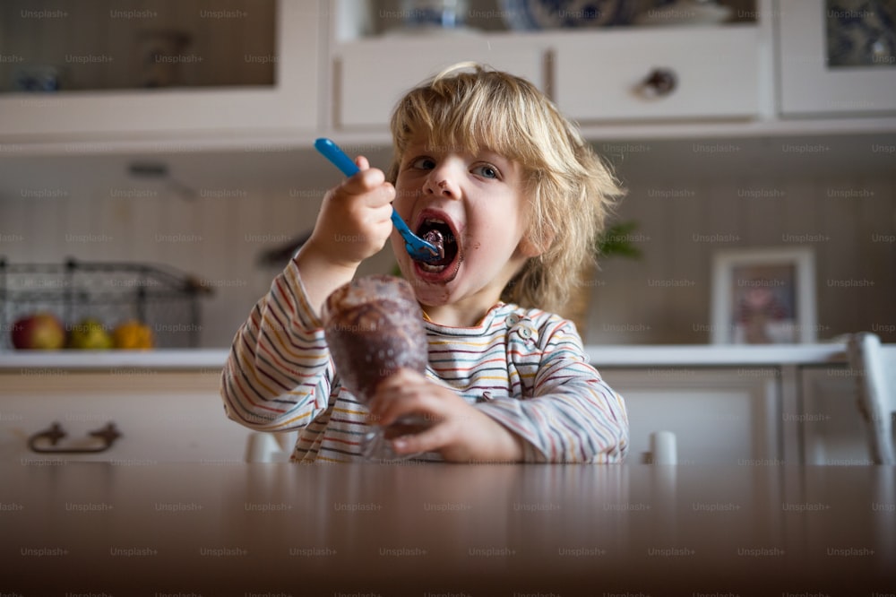 Retrato de menino pequeno com boca suja dentro de casa na cozinha em casa, comendo pudim.