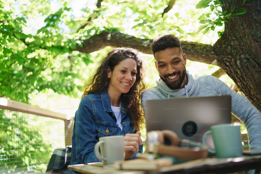 Una pareja feliz con una computadora portátil descansando al aire libre en una casa en el árbol, un fin de semana y un concepto de oficina remota.