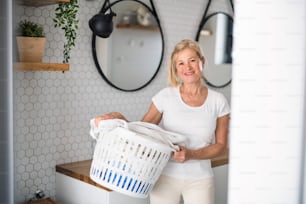Portrait of senior woman with laundry basket in bathroom indoors at home.