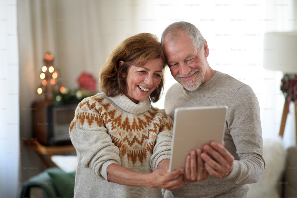 Front view of happy senior couple with tablet indoors at home at Christmas, taking selfie.