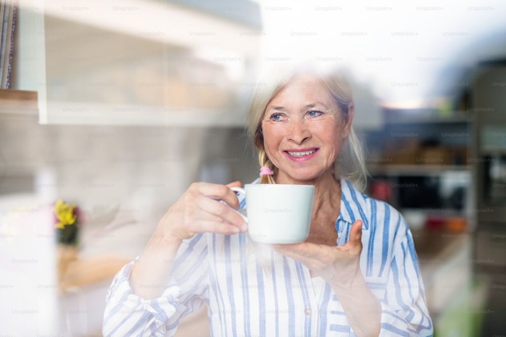 Portrait of senior woman with cup of coffee indoors at home. Shot through glass.