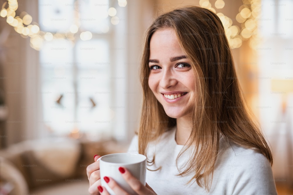 Retrato de una joven feliz en el interior de su casa en Navidad, sosteniendo una taza de café.
