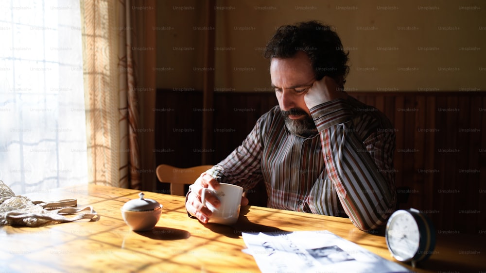 Portrait of unhappy poor mature man with coffee reading newspapers indoors at home, poverty concept.