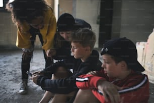 Front view of group of teenagers gang sitting indoors in abandoned building, using smartphones.