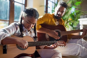 A happy father with small daughter indoors at home, playing guitar.