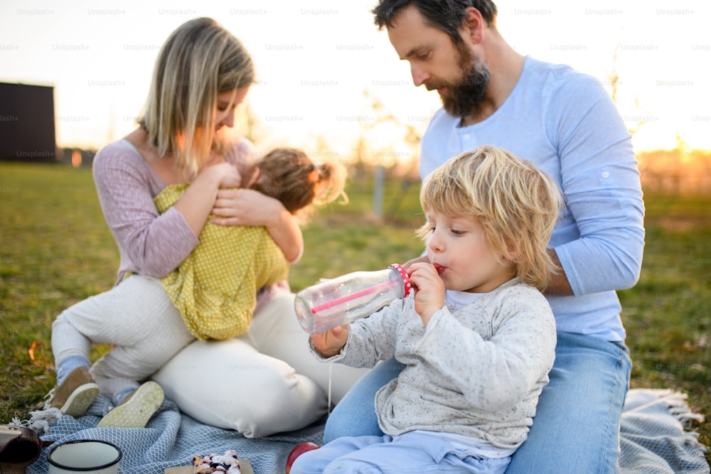 Front view of family with two small children having picnic outdoors in spring nature at sunset.