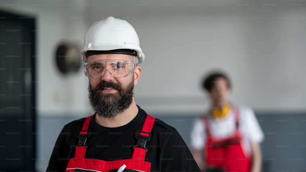 A portrait of worker with helmet and protective glasses indoors in factory looking at camera.