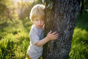 春の自然の中で屋外に立ち、木を抱きしめている小さな悲しい男の子の正面図。