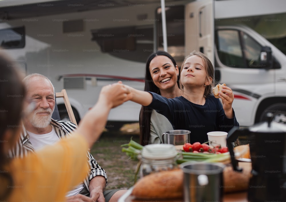 Une famille multigénérationnelle assise et mangeant à l’extérieur en voiture, voyage de vacances en caravane.