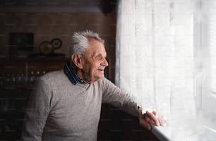 Ein Porträt eines älteren Mannes, der zu Hause drinnen steht und durch das Fenster schaut.