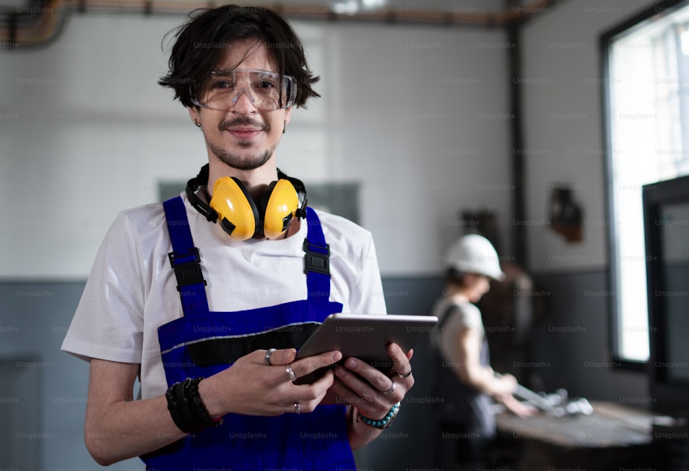 A portrait of young worker with tablet indoors in factory looking at camera.
