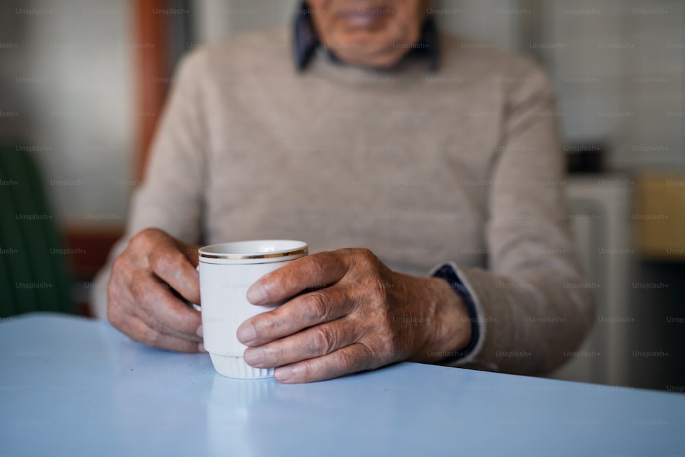 Un vieil homme méconnaissable avec une tasse de thé assis à l’intérieur à la maison, en train de se reposer.