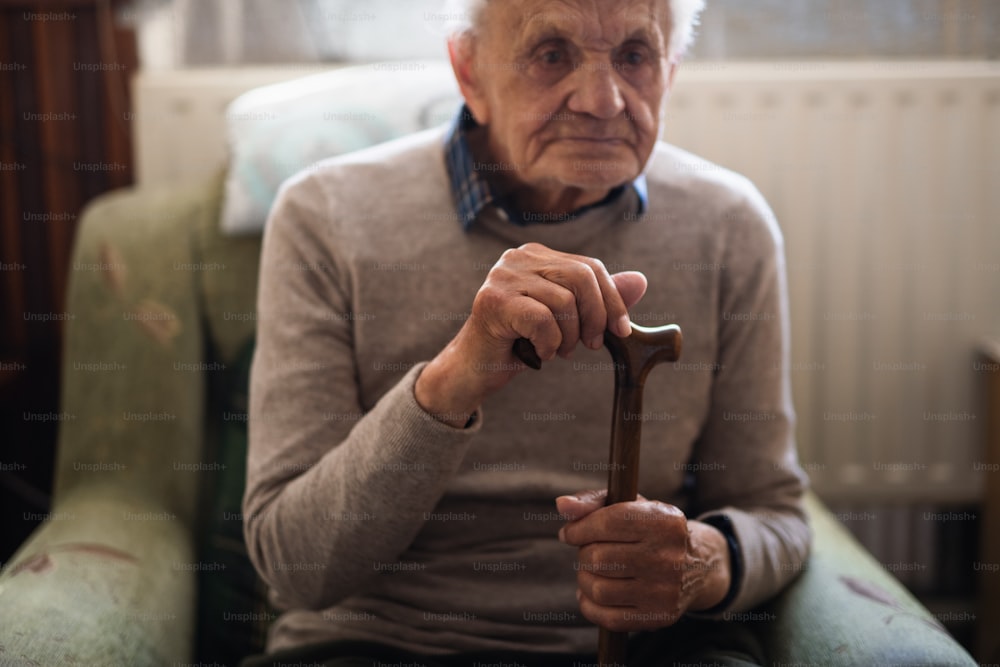 A sad elderly man with walking stick sitting on armchair indoors at home, resting.
