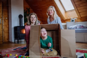 다운 증후군을 앓고있는 소년과 그의 어머니와 할머니가 집에서 함께 상자를 가지고 놀고 있습니다.