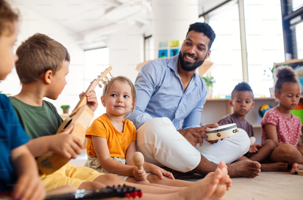 Gruppe kleiner Kindergartenkinder mit männlichem Lehrer, der drinnen im Klassenzimmer auf dem Boden sitzt und Musikinstrumente spielt.
