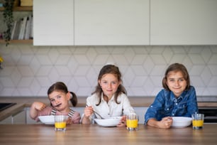 A portrait of three girls sisters indoors at home, looking at camera when eating breakfast.