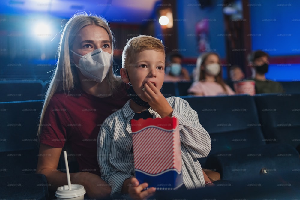 A small boy with mother eating popcorn in the cinema, watching movies and coronavirus concept.