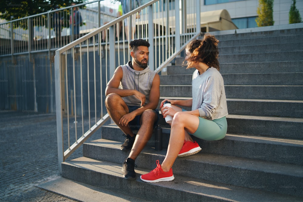 Un homme et une femme amis assis sur les escaliers à l’extérieur en ville, parlant après l’exercice.