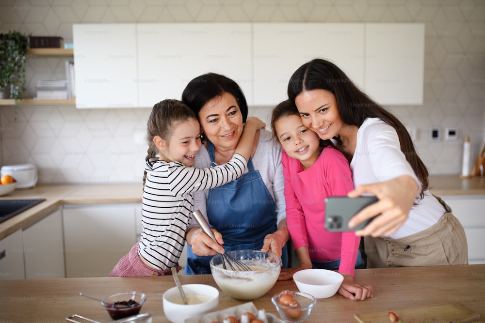 Happy small girls with a mother and grandmother making pancake mixture and taking selfie indoors at home