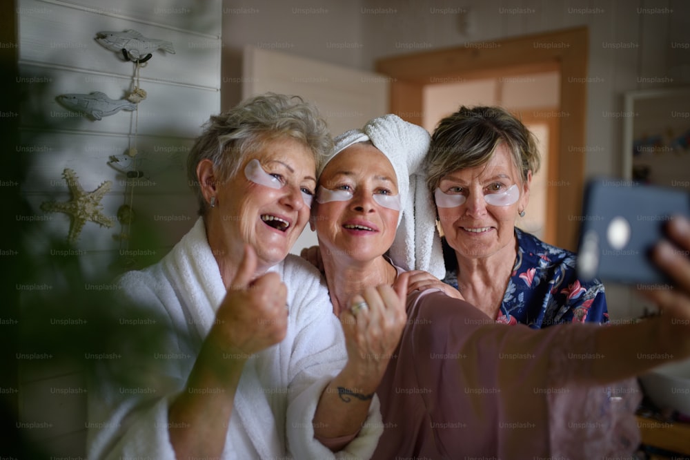 Happy senior women friends in bathrobes taking a selfie indoors at home, selfcare concept.